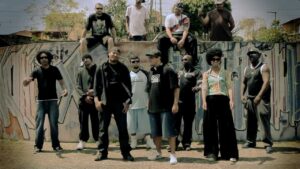 A cena do clipe da música 'Perigo', produzida pelo DJ Nato PK, registra uma reunião de peso que simboliza a força do coletivo e do selo Pau-de-Dá-em-Doido. Além do DJ, o quadro reúne os MCs André Sagat, Max Musicamente, Preto-R, Enézimo, Eri Q.i e Stefanie, que também emprestam suas vozes e líricas à faixa. Posicionados em formação de grupo à frente de um extenso muro com 'graffitis' e 'tags', os artistas reforçam a ocupação urbana e a estética clássica do hip hop de rua.