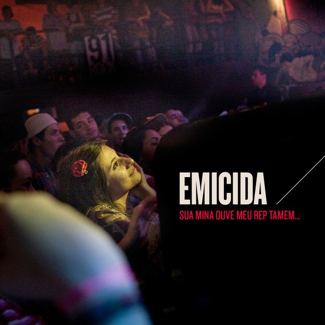 A capa do EP de Emicida, intitulado 'SUA MINA OUVE MEU REP TAMEM...', apresenta uma fotografia escura e granulada capturada em um show de rap em um ambiente noturno lotado. O visual central é o rosto de uma jovem fã, desfocada mas claramente visível, olhando para cima e para a direita (em direção ao palco) com uma expressão de admiração e conexão. Ela tem uma flor vermelha no cabelo e pulseiras neon em primeiro plano à esquerda. Atrás dela, a multidão está imersa na escuridão, com silhuetas e alguns rostos visíveis sob iluminação fraca. No canto inferior direito, o texto 'EMICIDA' está em letras maiúsculas brancas, em negrito, com uma linha branca diagonal sobreposta às letras finais, e abaixo, em vermelho, o título 'SUA MINA OUVE MEU REP TAMEM...' em texto maiúsculo.