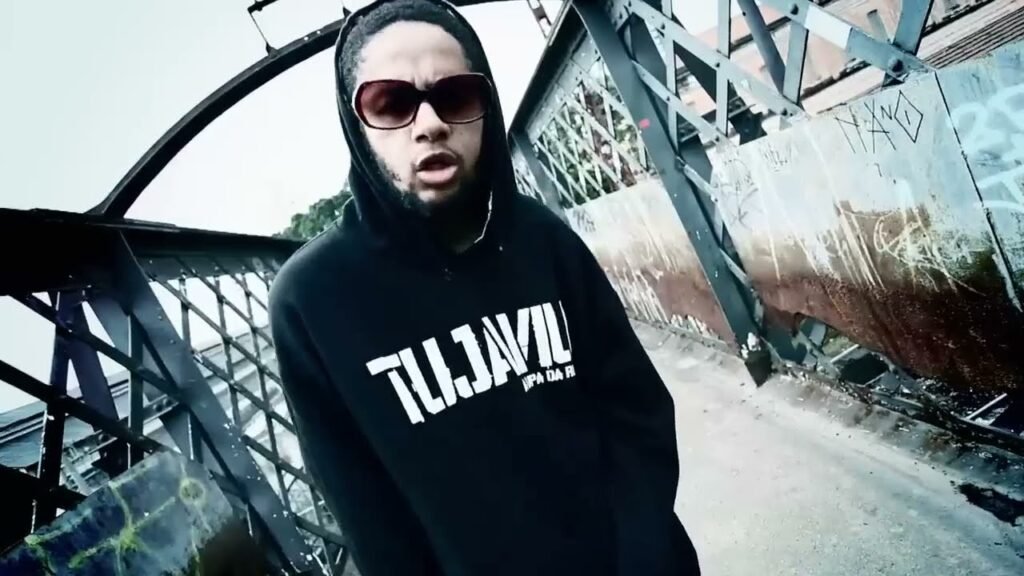 Emicida aparece no centro de uma passarela de ferro no clipe de 'Triunfo', trajando um moletom preto da marca 'TUJAVIU' e óculos escuros enquanto rima em meio a uma estética urbana industrial. O cenário, composto por grades metálicas e pichações, captura a crueza e a força do movimento independente no final dos anos 2000.