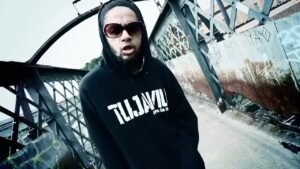 Emicida aparece no centro de uma passarela de ferro no clipe de 'Triunfo', trajando um moletom preto da marca 'TUJAVIU' e óculos escuros enquanto rima em meio a uma estética urbana industrial. O cenário, composto por grades metálicas e pichações, captura a crueza e a força do movimento independente no final dos anos 2000.