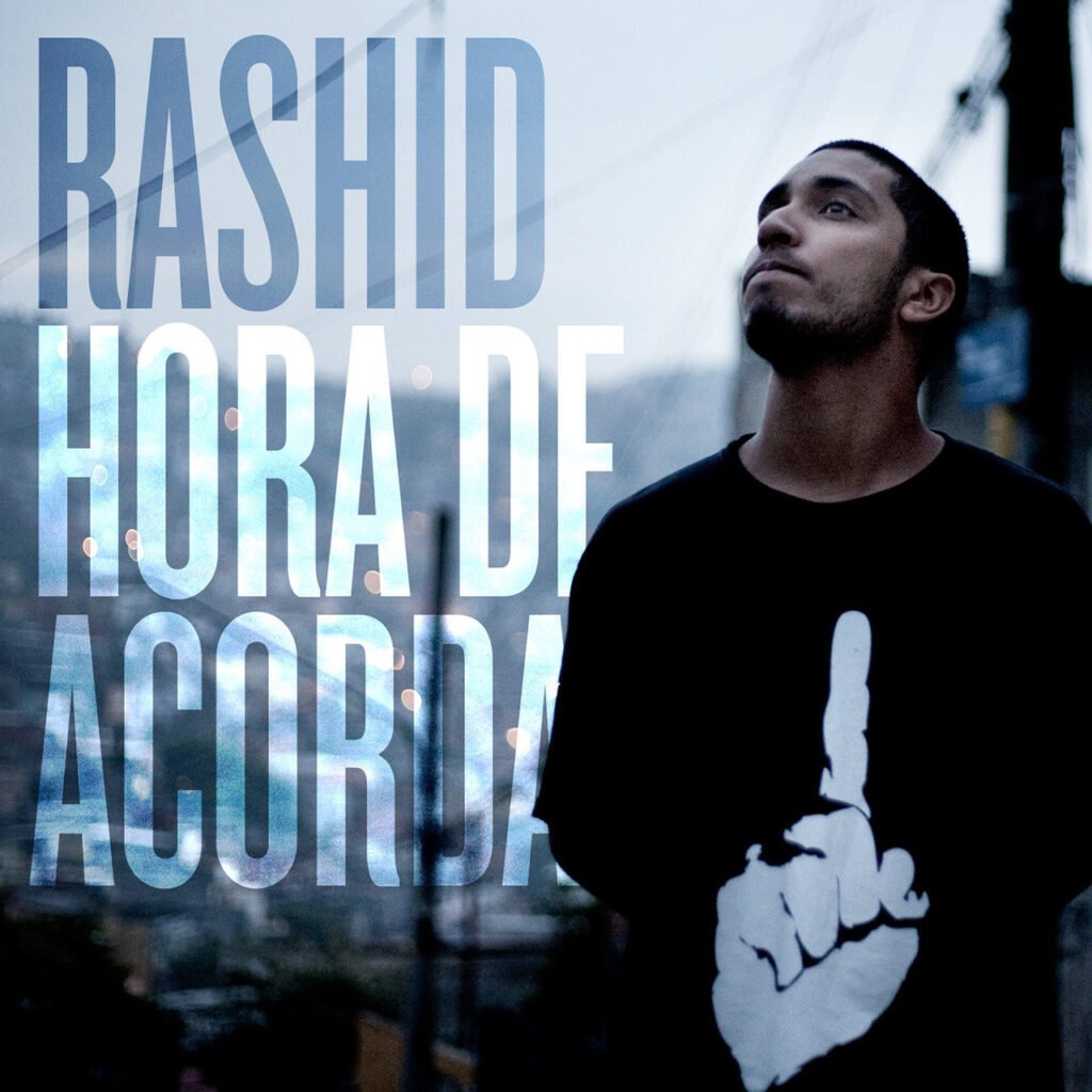 A capa da mixtape 'Hora de Acordar' exibe o rapper Rashid posicionado do lado direito da imagem, enquadrado do peito para cima, com o rosto virado para o alto. Ele veste uma camiseta preta estampada com a ilustração de uma mão branca com o dedo indicador apontado para cima. O fundo da imagem é desfocado e mostra uma paisagem urbana com casas amontoadas em um morro, postes, fios elétricos e luzes circulares em tons frios de azul e cinza. Ocupando a metade esquerda da composição, o texto com o nome 'RASHID' e o título 'HORA DE ACORDAR' é apresentado em três linhas, utilizando letras maiúsculas grandes, brancas e semitransparentes que se sobrepõem à paisagem ao fundo.