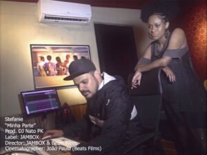 A cena do clipe de 'Minha Parte' registra o ambiente de criação no estúdio, onde o produtor DJ Nato PK opera o software de edição em frente a um monitor, vestindo jaqueta corta-vento preta e boné, enquanto a rapper Stefanie observa o processo apoiada no encosto de sua cadeira. O cenário é composto por paredes de tratamento acústico simples, um ar-condicionado e um quadro ao fundo que exibe uma fotografia artística de mulheres sentadas de costas, trazendo um ar de imersão técnica e profissionalismo. No canto inferior esquerdo, as informações de crédito confirmam a direção da JAMBOX & Beats Films, selando a estética de bastidores que caracteriza o registro dessa colaboração fundamental para o rap nacional.