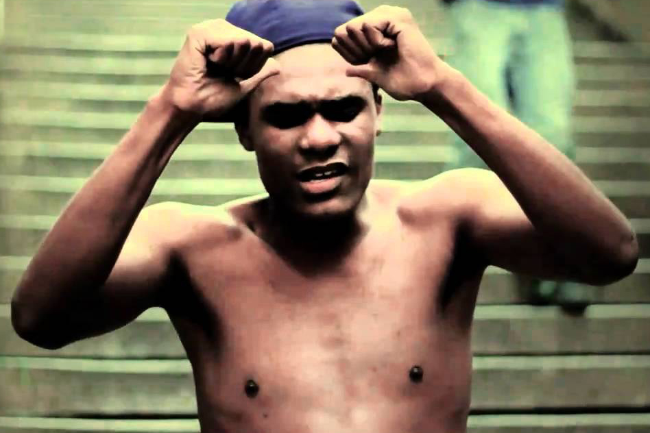 A cena do clipe 'Crises' exibe o cantor Terra Preta em primeiro plano do lado direito, enquadrado do peito para cima e com a boca entreaberta enquanto canta. Ele tem cabelo escuro e curto, veste uma camiseta preta, usa óculos escuros de armação espessa e uma grossa corrente de prata no pescoço. O cenário urbano noturno ao fundo encontra-se totalmente desfocado, revelando pontos de luz amarelada com efeito 'bokeh' provenientes da iluminação pública e a silhueta escura do que parece ser a estrutura de um viaduto ou ponte ao longe.