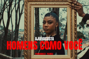 Uma cena do clipe 'Homens Como Você', da artista Ajulliacosta, apresenta a rapper centralizada dentro de uma moldura dourada ornamentada, que é segurada por mãos femininas com unhas longas e anéis dourados. Ajulliacosta encara a câmera com uma expressão séria, usando argolas grandes e o cabelo preso em um rabo de cavalo alto. O fundo mostra uma paisagem urbana com árvores e casas sob uma luz diurna suave. Sobreposto à imagem, o nome 'AJULIACOSTA' aparece em branco e o título 'HOMENS COMO VOCÊ' em letras garrafais vermelhas, com o crédito 'FERNANDEZZ FILMES' logo abaixo.