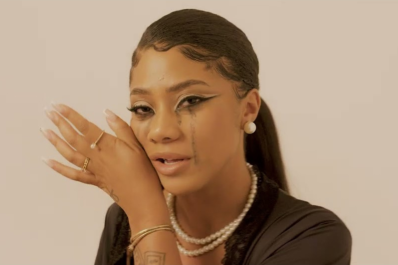 Imagem do clipe 'Não Foi do Nada', da artista Ajulliacosta, mostra um retrato da rapper em close-up contra um fundo claro. Ela aparece com uma expressão vulnerável, com lágrimas escuras de maquiagem escorrendo pelos olhos e uma mão próxima ao rosto, revelando anéis e unhas longas. Ajulliacosta usa o cabelo preso em um rabo de cavalo polido com baby hair estilizado, além de acessórios clássicos como brincos de pérola e um colar de pérolas de duas voltas sobre uma roupa escura com detalhes em renda. A iluminação é suave, destacando os detalhes da maquiagem e das joias