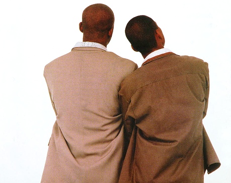 Fotografia de Claudinho e Buchecha de costas contra um fundo branco liso. Claudinho, à esquerda, veste um paletó bege claro sobre uma camisa branca com colarinho texturizado, enquanto Buchecha, à direita, veste um paletó marrom escuro, também sobre uma camisa branca. Ambos têm a cabeça raspada ou cabelo bem curto, estão posicionados próximos um do outro com os braços aparentemente cruzados à frente, e com as cabeças inclinadas como se estivessem olhando para algo acima e à direita.