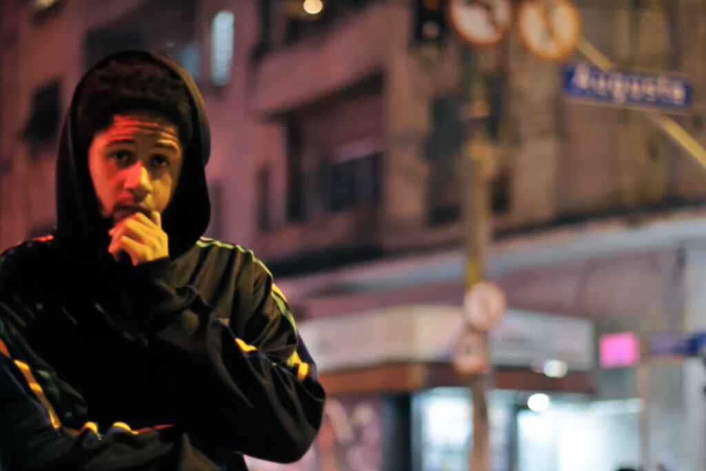 Cena noturna do videoclipe Rua Augusta mostrando o rapper Emicida em primeiro plano, vestindo uma jaqueta escura com o capuz levantado e detalhes coloridos nas mangas. Ele olha de forma pensativa para a câmera com uma das mãos apoiada no queixo, tendo ao fundo um cenário urbano urbano desfocado iluminado por luzes quentes, onde é possível identificar a inscrição 'Augusta' em uma placa azul de sinalização de rua.