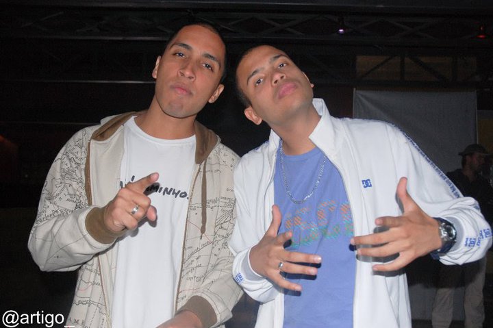 Fotografia dos rappers Rashid, à esquerda, e Projota, à direita, posando juntos em um ambiente interno com pouca iluminação. Rashid veste uma camiseta branca sob um moletom claro aberto com estampa de mapa e aponta o dedo indicador direito em direção à câmera. Projota usa uma camiseta azul com uma corrente prateada e uma jaqueta branca, fazendo gestos soltos com as duas mãos. No canto inferior esquerdo da imagem, há uma marca d'água com o texto '@artigo'.