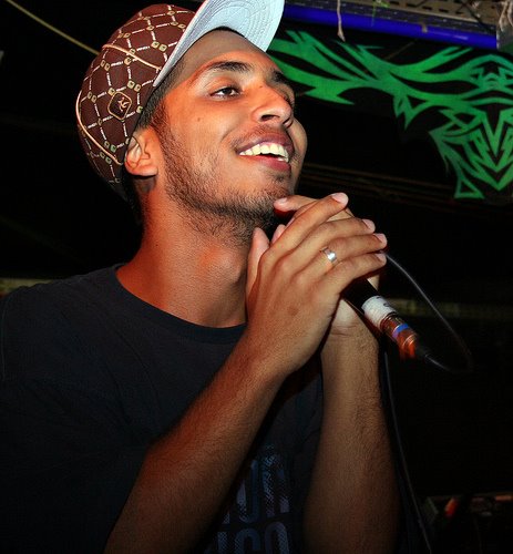 Fotografia do rapper Rashid sorrindo e segurando um microfone com as duas mãos perto do queixo. Ele tem barba rala e veste uma camiseta escura com um boné marrom de aba reta e estampa geométrica, posicionado contra um fundo escuro que exibe um detalhe de arte urbana verde brilhante.