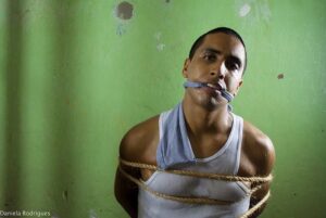 O rapper Rashid aparece em primeiro plano, com o corpo suado e uma expressão de resistência, posicionado contra uma parede verde desgastada. Ele está amarrado com cordas de sisal que circundam seus braços e peito, e possui uma mordaça feita de tecido azul claro presa em sua boca. Vestindo uma regata branca de ribana, ele encara diretamente a câmera com a cabeça levemente inclinada, em uma cena dramática e intensa extraída do clipe 'Vou Ser Mais'.