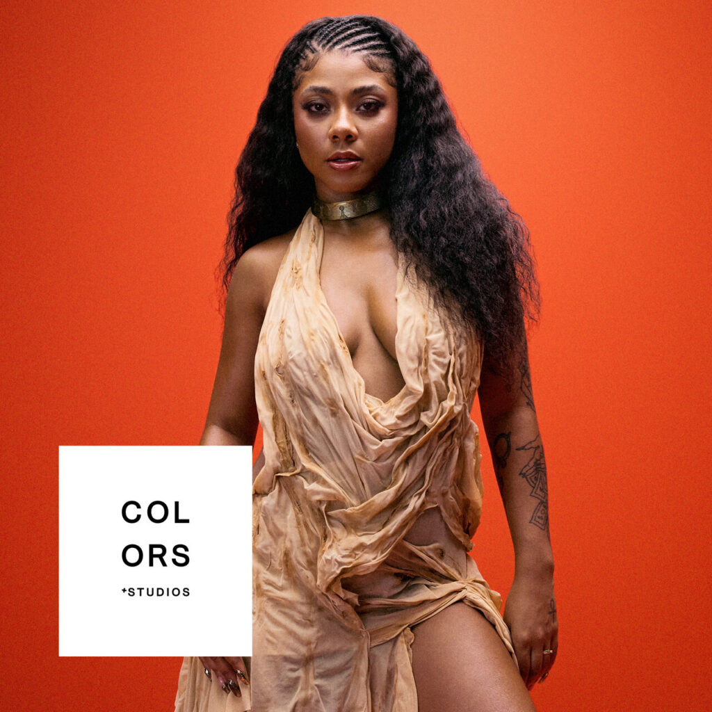 A imagem apresenta a artista Ajulliacosta centralizada contra um fundo laranja sólido. Ela está de pé, vestindo um vestido de tecido bege com textura amassada e drapeada, que possui diversas aberturas e rasgos propositais. Seu cabelo é longo, preto e ondulado, com tranças rasteiras (cornrows) no topo da cabeça. No pescoço, ela usa uma gargantilha metálica dourada. No canto inferior esquerdo, há um quadrado branco com o texto 'COLORS + STUDIOS' em letras pretas.