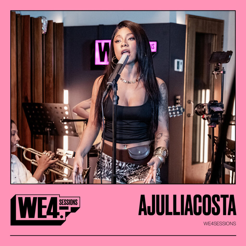 A capa de 'WE4SESSIONS' destaca a artista Ajulliacosta em um momento de performance dentro de um estúdio de gravação, emoldurada por uma borda rosa vibrante na parte inferior. No centro da imagem, ela aparece cantando em um microfone de pedestal, usando uma regata preta, shorts com estampa animal print, um cinto marrom com pequena pochete acoplada e diversos acessórios prateados e dourados. O cenário ao fundo revela a atmosfera de uma sessão ao vivo, com a presença parcial de músicos — uma trompetista à esquerda e um violonista atrás —, além de equipamentos de filmagem e painéis acústicos de madeira. O design da peça é finalizado com o logotipo 'WE4 SESSIONS' e o nome da artista em letras pretas robustas sobre o fundo rosa, conferindo um visual moderno e dinâmico que remete a um registro íntimo e profissional.