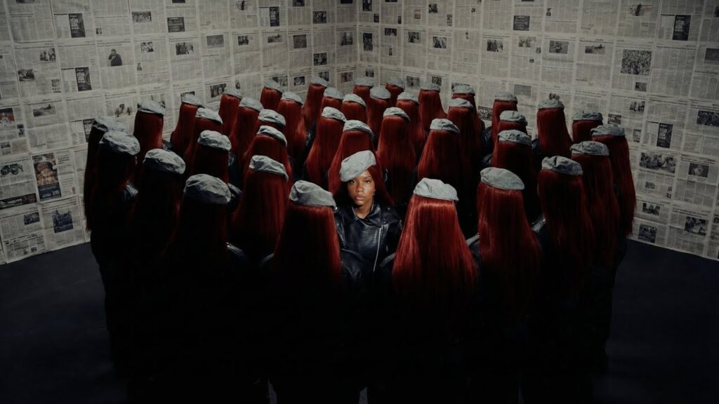 Cena do videoclipe 'BPP' da rapper Majis. No centro da imagem, Majis, uma mulher negra, está de pé olhando diretamente para a câmera com uma expressão séria. Ela veste uma jaqueta de couro preta e uma boina cinza inclinada na cabeça. Ela está cercada por uma multidão de mulheres que estão de costas para a câmera; todas usam jaquetas pretas, boinas cinzas idênticas e têm longos cabelos lisos de cor vermelho-escuro, criando um efeito de replicação. O cenário é o canto de uma sala onde as paredes estão completamente cobertas por folhas de jornal antigas com textos e fotos em preto e branco.