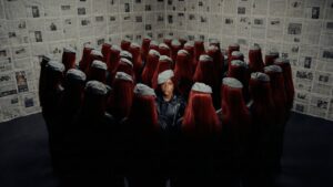 Cena do videoclipe 'BPP' da rapper Majis. No centro da imagem, Majis, uma mulher negra, está de pé olhando diretamente para a câmera com uma expressão séria. Ela veste uma jaqueta de couro preta e uma boina cinza inclinada na cabeça. Ela está cercada por uma multidão de mulheres que estão de costas para a câmera; todas usam jaquetas pretas, boinas cinzas idênticas e têm longos cabelos lisos de cor vermelho-escuro, criando um efeito de replicação. O cenário é o canto de uma sala onde as paredes estão completamente cobertas por folhas de jornal antigas com textos e fotos em preto e branco.