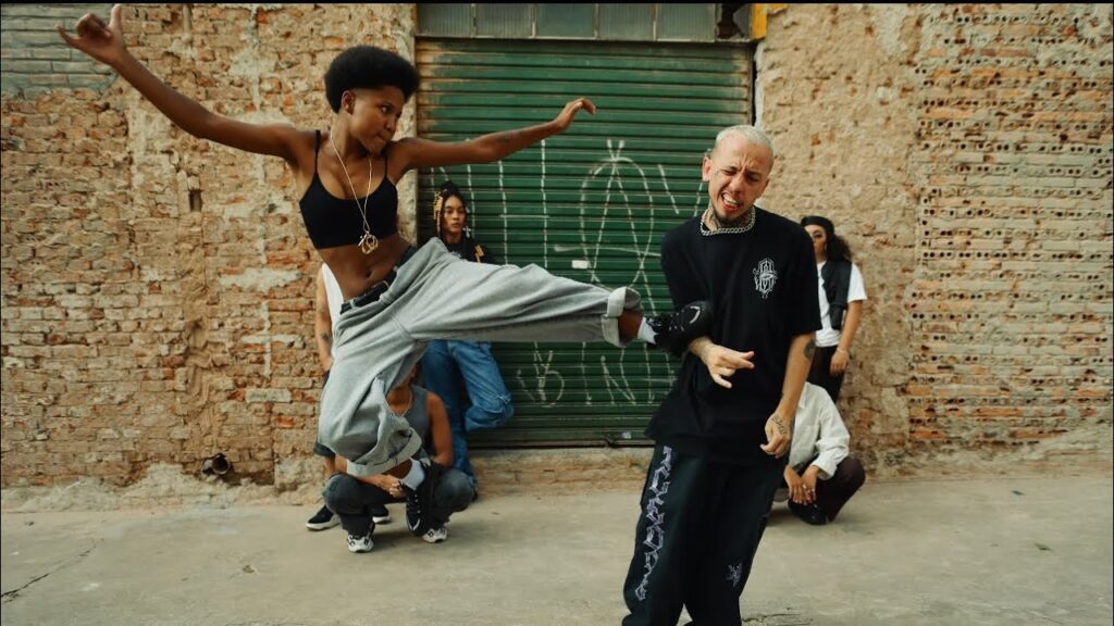 Cena do videoclipe 'Fora da Caixa' da rapper Majis. No centro da imagem, Majis, uma mulher negra com cabelo curto estilo crespo/black power, aparece em movimento dinâmico, saltando no ar enquanto desfere um chute lateral com a perna esquerda estendida. Ela veste um top preto, calça de moletom cinza larga e tênis pretos, exibindo um colar comprido com pingente dourado. O golpe atinge a costela de um homem branco de cabelo raspado e descolorido, que reage com uma careta de dor; ele veste uma camiseta preta larga, calça preta com estampas brancas na perna e uma corrente prateada no pescoço. Ao fundo, outras pessoas observam a cena em um ambiente urbano, com uma grande porta de metal verde e paredes de tijolos expostos e desgastados.