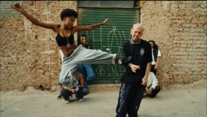 Cena do videoclipe 'Fora da Caixa' da rapper Majis. No centro da imagem, Majis, uma mulher negra com cabelo curto estilo crespo/black power, aparece em movimento dinâmico, saltando no ar enquanto desfere um chute lateral com a perna esquerda estendida. Ela veste um top preto, calça de moletom cinza larga e tênis pretos, exibindo um colar comprido com pingente dourado. O golpe atinge a costela de um homem branco de cabelo raspado e descolorido, que reage com uma careta de dor; ele veste uma camiseta preta larga, calça preta com estampas brancas na perna e uma corrente prateada no pescoço. Ao fundo, outras pessoas observam a cena em um ambiente urbano, com uma grande porta de metal verde e paredes de tijolos expostos e desgastados.