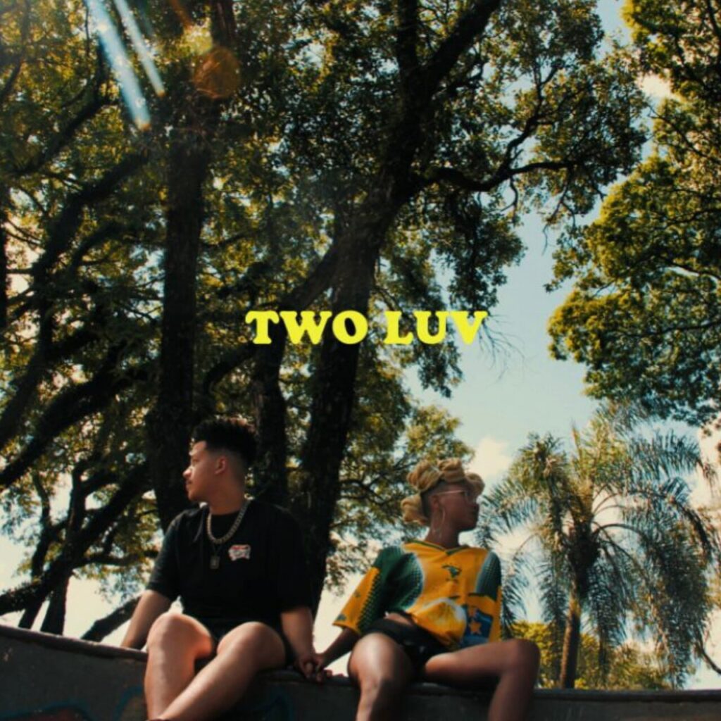 A capa do EP 'TWO LUV', de Majis e RXDS, exibe uma fotografia ao ar livre focada em um casal sentado de costas um para o outro, mas de mãos dadas, sobre uma mureta de concreto. O rapaz à esquerda veste camiseta preta e corrente prateada, enquanto a jovem à direita usa uma camisa amarela e verde e óculos. O cenário ao fundo é composto por árvores altas e folhagens sob o céu claro, e o título 'TWO LUV' aparece centralizado em letras amarelas em bloco.
