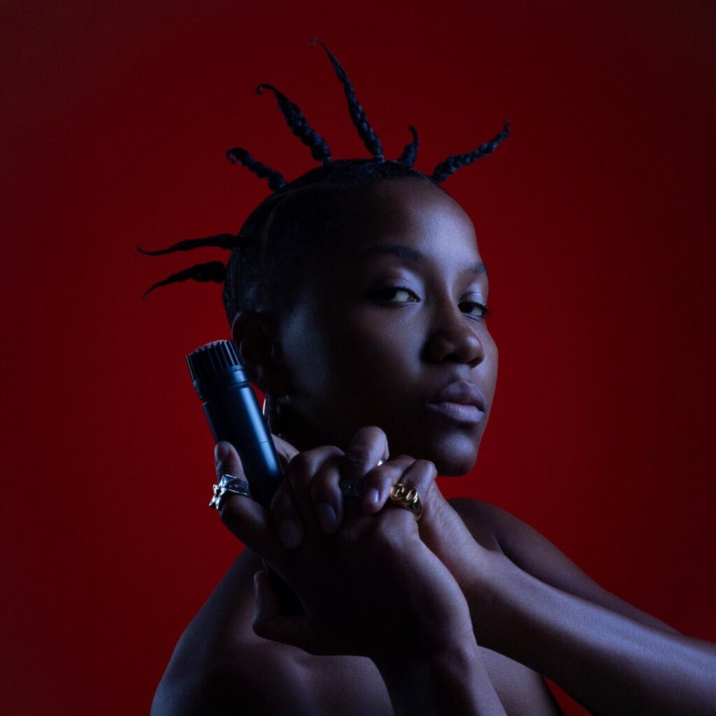 Capa do single 'Fora da Caixa' da rapper Majis. Na imagem, Majis é vista de perfil com o rosto virado para a câmera, com uma expressão séria e imponente. Ela é uma mulher negra, com os ombros nus e o cabelo estilizado em pequenas tranças espetadas para cima, lembrando o estilo de penteado Bantu knots ou espetos estilizados que formam uma coroa ao redor de sua cabeça. A iluminação é dramática e focada, vinda da lateral, destacando os contornos de seu rosto e braços contra um fundo vermelho-escuro e sólido. Suas mãos estão cruzadas à frente do corpo, na altura do queixo, segurando um microfone de estúdio preto. Ela usa vários anéis chamativos nos dedos, incluindo um anel prateado no dedão e um anel dourado no dedo médio.