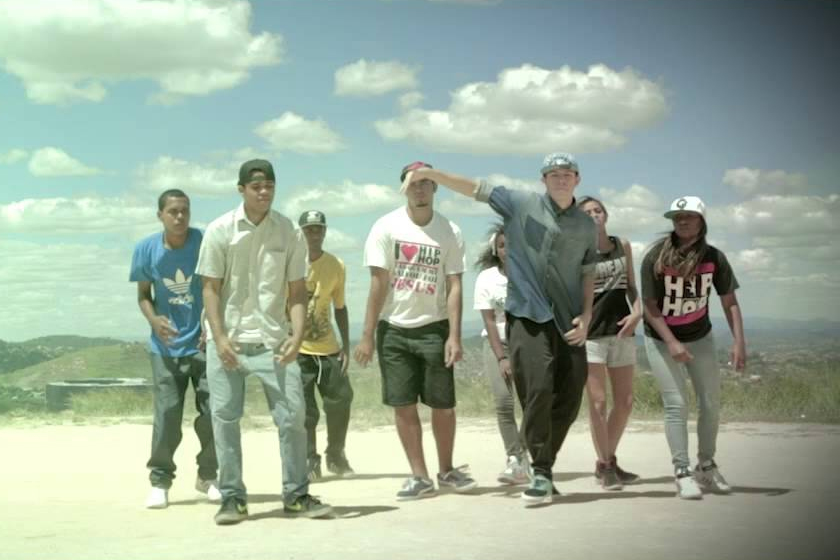 A imagem mostra um grupo de oito jovens, homens e mulheres, em uma cena externa do clipe 'Anônima' de Tamara Franklin. Eles estão organizados em uma formação de dança sobre um terreno aberto, vestindo roupas típicas da cultura hip-hop, como bonés e camisetas estampadas, sob um céu aberto e iluminado. A fotografia captura um momento de movimento coordenado, com uma paisagem de colinas ao horizonte, reforçando a estética urbana e comunitária do vídeo.