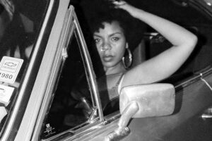 Trata-se de uma fotografia em preto e branco com granulação visível. A artista Tamara Franklin é vista através da janela de um carro antigo. Ela possui cabelo estilo afro e usa brincos de argola grandes. Seu braço esquerdo está flexionado com a mão tocando o topo da cabeça, enquanto o braço direito está apoiado na porta do veículo. O espelho retrovisor retangular cromado está em destaque no primeiro plano. No vidro lateral esquerdo, há adesivos, incluindo um com o logo da Chevrolet e o ano '1980'.