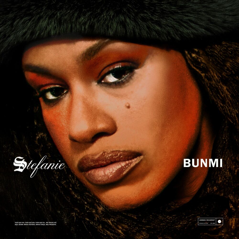 A capa do álbum BUNMI, da rapper Stefanie, apresenta um retrato em close-up da artista com olhar firme em direção à câmera. Ela usa maquiagem marcante nos olhos com sombra avermelhada, delineado preto e batom gloss. Na cabeça, veste um chapéu de pelúcia preto e o fundo da imagem é escuro. No canto inferior esquerdo, o nome 'Stefanie' está grafado em fonte cursiva estilo gótica na cor branca. Logo abaixo, lê-se em letras menores: 'TUDO QUE VIVI, TUDO QUE SOU, TUDO QUE FUI... ME TROUXE ATÉ AQUI. BUNMI: MINHA HISTÓRIA, MINHA FORÇA, MEU PRESENTE.' No canto inferior direito, destaca-se o título do álbum, 'BUNMI', em caixa alta e fonte sem serifa branca, acompanhado do logotipo retangular da gravadora 'JAMBOX RECORDS' na extremidade inferior.