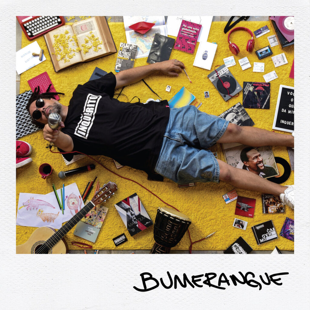 A capa do álbum Bumerangue, do grupo de rap Inquérito, é emoldurada por uma borda branca espessa no estilo de uma foto instantânea Polaroid, vista inteiramente de um ângulo superior (flat lay). No centro, o rapper Renan aparece deitado de costas sobre um tapete amarelo felpudo. Ele veste uma camiseta preta com a palavra 'INQUÉRITO' estampada no peito, bermuda jeans e óculos escuros, ostentando dreads no cabelo. Na mão direita levantada, ele segura um microfone de estilo vintage prateado. Ao redor do artista, espalhados em composição minuciosa sobre o tapete amarelo, estão diversos objetos pessoais e referências artísticas: um violão de náilon, um instrumento de percussão (djembe), livros, capas de discos de vinil (como do cantor Marvin Gaye), uma máquina de escrever antiga, fitas cassete, fones de ouvido vermelhos, desenhos infantis e pequenos cartões. Na borda branca inferior, o título do álbum, BUMERANGUE, aparece escrito em letras maiúsculas pretas em estilo manuscrito.