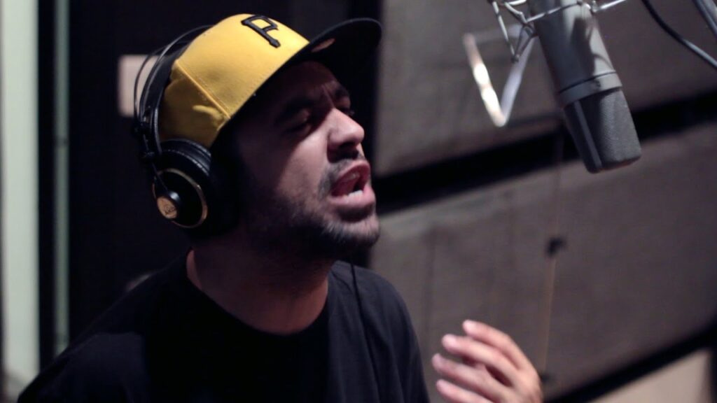 Fotografia extraída do clipe da música 'PÓ ESIA', do grupo Inquérito, focada no rapper Renan dentro de um estúdio de gravação. O artista aparece em plano médio, ligeiramente inclinado para a frente e de olhos fechados com uma expressão concentrada enquanto canta. Ele veste uma camiseta preta e usa fones de ouvidos pretos sobre um boné amarelo com aba preta que tem a letra 'P' bordada. No canto superior direito, sobressai o microfone de estúdio prateado fixado num suporte, para onde o artista direciona a voz com a mão direita parcialmente visível na parte inferior da imagem. O fundo do estúdio é escuro e com tratamento acústico.