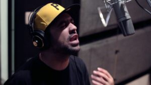 Fotografia extraída do clipe da música 'PÓ ESIA', do grupo Inquérito, focada no rapper Renan dentro de um estúdio de gravação. O artista aparece em plano médio, ligeiramente inclinado para a frente e de olhos fechados com uma expressão concentrada enquanto canta. Ele veste uma camiseta preta e usa fones de ouvidos pretos sobre um boné amarelo com aba preta que tem a letra 'P' bordada. No canto superior direito, sobressai o microfone de estúdio prateado fixado num suporte, para onde o artista direciona a voz com a mão direita parcialmente visível na parte inferior da imagem. O fundo do estúdio é escuro e com tratamento acústico.