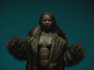 A imagem extraída do clipe música 'P.I.T.T.Y (Parecendo Uma Cafetina)' traz um enquadramento de meio-corpo da artista NandaTsunami, centralizada contra um fundo de tom azul-petróleo ou verde-azulado escuro. Ela aparece vestindo um casaco de pele volumoso e felpudo em tons de castanho e marrom sobre o peito entreaberto. Seu cabelo está penteado em tranças nagô com detalhes em tom de castanho-claro, e ela usa uma corrente dourada no pescoço, brincos, um piercing no umbigo e possui uma tatuagem na região do peito. A artista segura as bordas do casaco com ambas as mãos e olha diretamente para a câmera com expressão séria.
