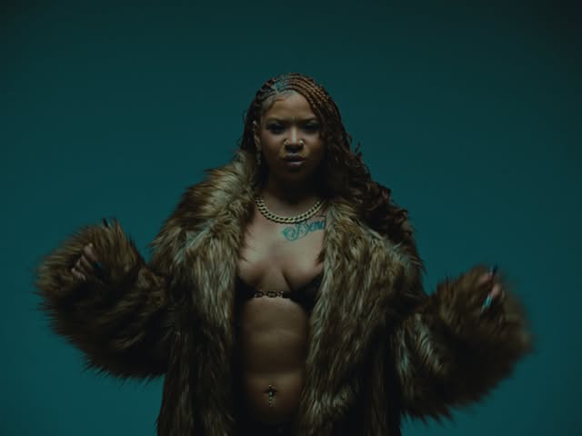 A imagem extraída do clipe música 'P.I.T.T.Y (Parecendo Uma Cafetina)' traz um enquadramento de meio-corpo da artista NandaTsunami, centralizada contra um fundo de tom azul-petróleo ou verde-azulado escuro. Ela aparece vestindo um casaco de pele volumoso e felpudo em tons de castanho e marrom sobre o peito entreaberto. Seu cabelo está penteado em tranças nagô com detalhes em tom de castanho-claro, e ela usa uma corrente dourada no pescoço, brincos, um piercing no umbigo e possui uma tatuagem na região do peito. A artista segura as bordas do casaco com ambas as mãos e olha diretamente para a câmera com expressão séria.