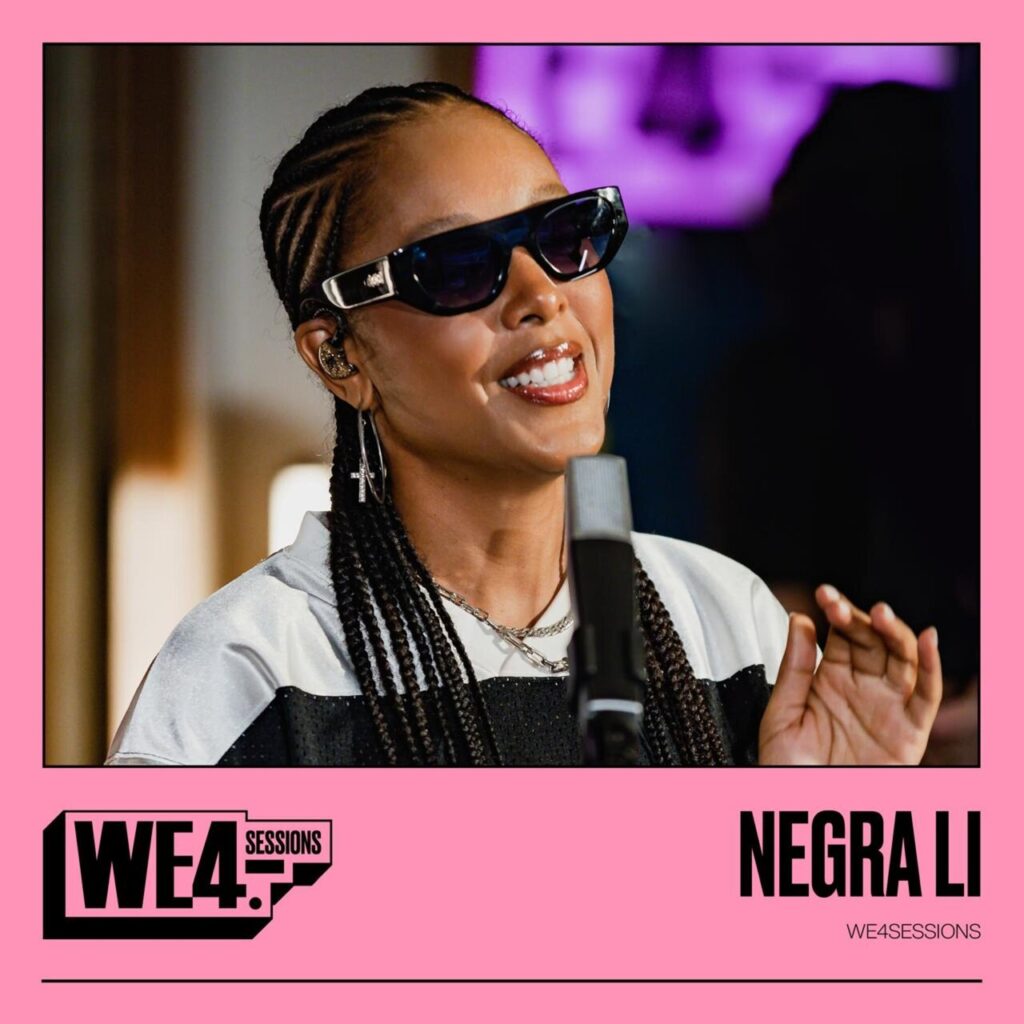 A capa do ep 'WE4SESSIONS' da cantora Negra Li é dividida em duas seções dentro de uma moldura cor-de-rosa. Na parte superior, uma foto mostra a artista sorrindo e olhando ligeiramente para cima em um ambiente iluminado. Ela usa óculos de sol pretos com hastes detalhadas em prata, tranças nagô na cabeça, brincos de argola, correntes no pescoço e um moletom esportivo preto e branco diante de um microfone de estúdio. A parte inferior tem fundo rosa. À esquerda, destaca-se o logotipo 'WE4 SESSIONS' em caixa preta e branca. À direita, o nome 'NEGRA LI' aparece em letras maiúsculas pretas e chamativas, acompanhado abaixo da palavra 'WE4SESSIONS' em fonte menor.