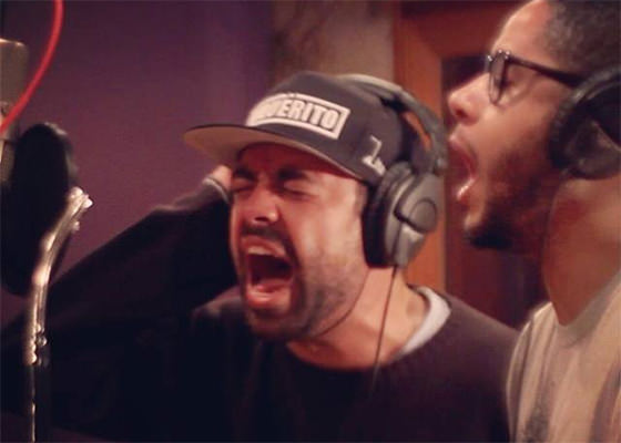 Fotografia em estúdio de gravação que mostra os rappers Renan (do grupo Inquérito) e Emicida cantando diante do microfone. Ao centro, Renan aparece em plano médio, de olhos fechados e boca bem aberta em um gesto expressivo e intenso de canto. Ele veste um moletom preto, usa fones de ouvido grandes sobre um boné escuro com a palavra 'INQUÉRITO' estampada em branco na parte frontal e segura os fones com a mão direita. À direita, em segundo plano e em perfil, Emicida surge também com a boca aberta cantando, usando óculos de armação preta e fones de ouvido. No canto esquerdo da imagem, destaca-se a estrutura prateada do microfone de estúdio com o pop filter preto posicionado à frente dos artistas. A iluminação do estúdio é quente e intimista.