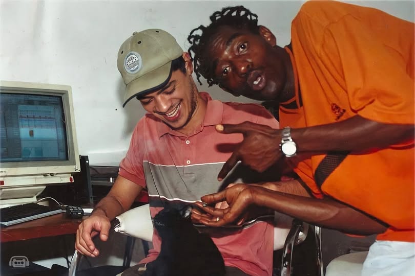 Fotografia no estúdio mostrando o produtor Daniel Ganjaman e o rapper Sabotage. À esquerda, Daniel Ganjaman está sentado diante de um monitor de computador antigo, usando boné bege e polo com faixas pretas e cinzas. Ele sorri enquanto acaricia o gato preto no seu colo. À direita, Sabotage aparece de pé, curvado em direção a Daniel, vestindo uma camiseta alaranjada e exibindo cabelo com tranças curtas. Ele aponta com a mão direita para o gato enquanto estende a mão esquerda, olhando expressivamente para a câmera. Ao fundo, vê-se a parede clara do estúdio e equipamentos.