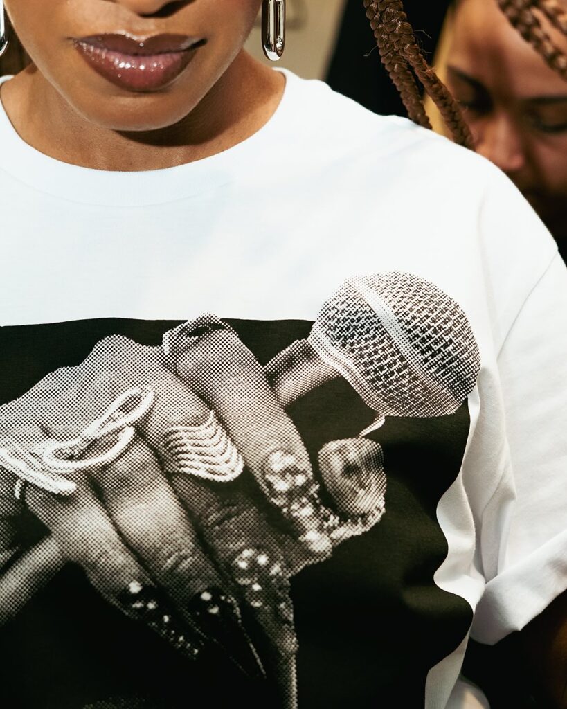 A imagem mostra um enquadramento aproximado em detalhe do peito de uma mulher usando uma camiseta branca. Na estampa da camiseta, há uma foto em tom meio-tom (reticulada) em preto e branco da mão de uma mulher segurando um microfone. Ela exibe unhas compridas pintadas de escuro com detalhes brilhantes e usa anéis prateados marcantes nos dedos, incluindo um anel largo e trabalhado. Na parte superior da imagem, aparece parcialmente o rosto da mulher que veste a camiseta, destacando seus lábios com gloss, brincos prateados pendentes e tranças no cabelo.