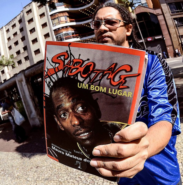 O escritor Toni C., autor da biografia, segura em primeiro plano o livro 'Sabotage: Um Bom Lugar'. Ele é um homem de pele negra com cabelo trançado, que usa óculos de sol de armação fina prateada e uma camisola desportiva azul e preta. A capa do livro, apresentada em perspetiva próxima da câmara, exibe em destaque o rosto em tom sério do rapper Sabotage em plano fechado. Na parte superior da capa, o nome 'SABOTAGE' surge numa fonte vermelha estilizada com contorno preto. Logo abaixo, lê-se o subtítulo 'UM BOM LUGAR' em letras maiúsculas amarelas. No canto inferior esquerdo do livro, encontram-se as inscrições 'BIOGRAFIA OFICIAL DE Toni C.' e 'Mauro Mateus do...'. Ao fundo da imagem, desfocada pela profundidade de campo, avista-se uma rua urbana com edifícios de arquitetura clássica.