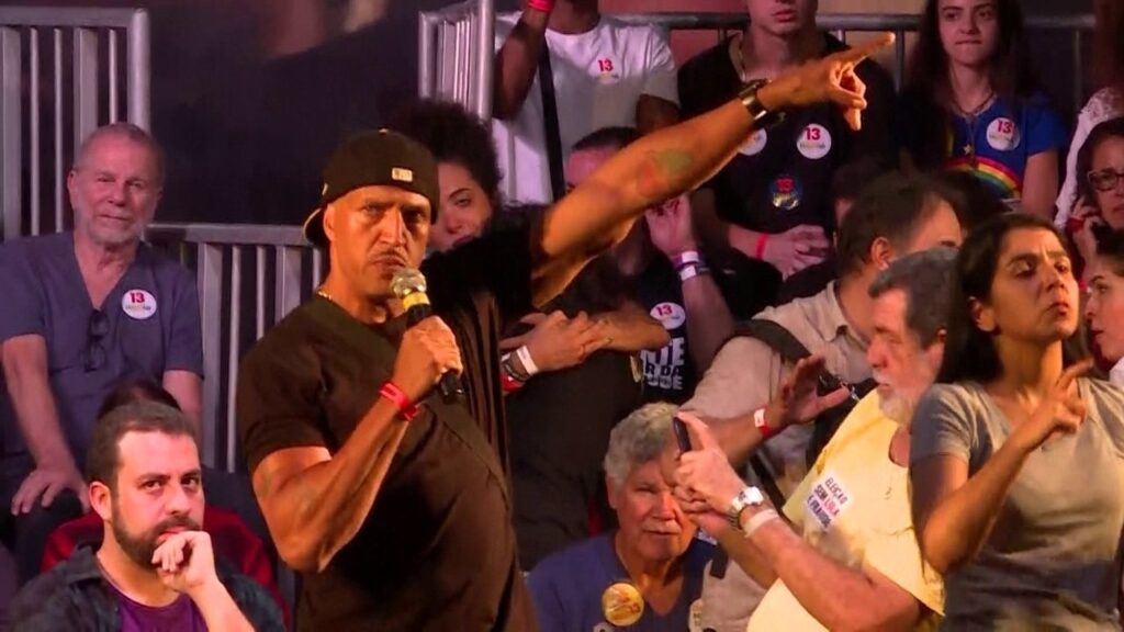 Fotografia capturada durante um comício político onde o rapper Mano Brown discursa ao microfone num ambiente de palco. Em primeiro plano, do lado esquerdo, Mano Brown aparece em plano médio, vestindo uma camiseta preta, correntes ao pescoço e um boné preto virado para trás. Com uma expressão séria e assertiva, segura um microfone preto com detalhes em dourado na mão direita e estende o braço esquerdo na diagonal para cima, gesticulando com o indicador apontado. Ao seu redor, uma plateia diversificada acompanha o momento. Em baixo, no canto esquerdo, destaca-se Guilherme Boulos de barba e camiseta roxa ouvindo atentamente. Do lado direito, em primeiro plano, uma mulher faz tradução em LIBRAS (Língua Brasileira de Sinais) e, ao seu lado, um homem com camiseta amarela onde se lê